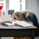 Pessoa exausta com a cabeça apoiada na mesa, cercada por livros e cadernos, simbolizando burnout e sobrecarga emocional.