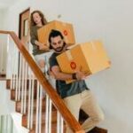 Casal carregando caixas juntos em uma escada, simbolizando cooperação e trabalho em equipe no relacionamento.