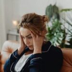 Mulher sentada no sofá com as mãos cobrindo o rosto, simbolizando o impacto emocional do estresse.