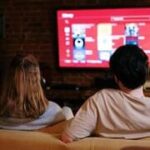 Casal sentado em um sofá assistindo televisão, representando rotina e desgaste no relacionamento.