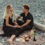 Casal sentado em um cobertor na praia, com um piquenique ao redor, representando rotina e desgaste no relacionamento.