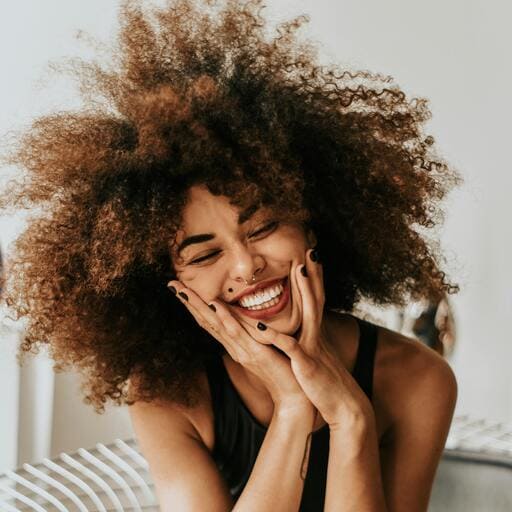 Pessoa com cabelo volumoso e cacheado, sentada com as mãos apoiadas no rosto, simbolizando felicidade e bem-estar.